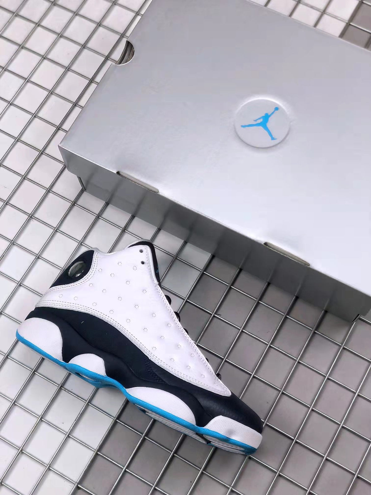 Air Jordan 13 sneaker beside a light-colored shoebox, showing the white perforated upper, black midsole, and blue outsole accent.