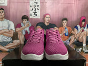 Front view of pink Nike Zoom basketball sneakers with textured uppers, white midsoles, and pink accents, displayed against an illustrated basketball backdrop.