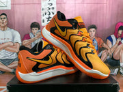 Angled view of the orange and black sneakers displaying the side branding, laces, heel lettering, and contrasting outsole.