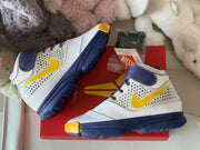 Angled top-down view of the pair on a red shoebox, showing the white and purple construction, yellow accents, and textured soles.