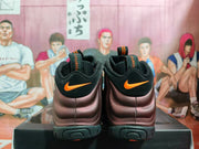 Rear view of brown and black Nike Foamposite-style sneakers with orange heel logos on a black shoebox.