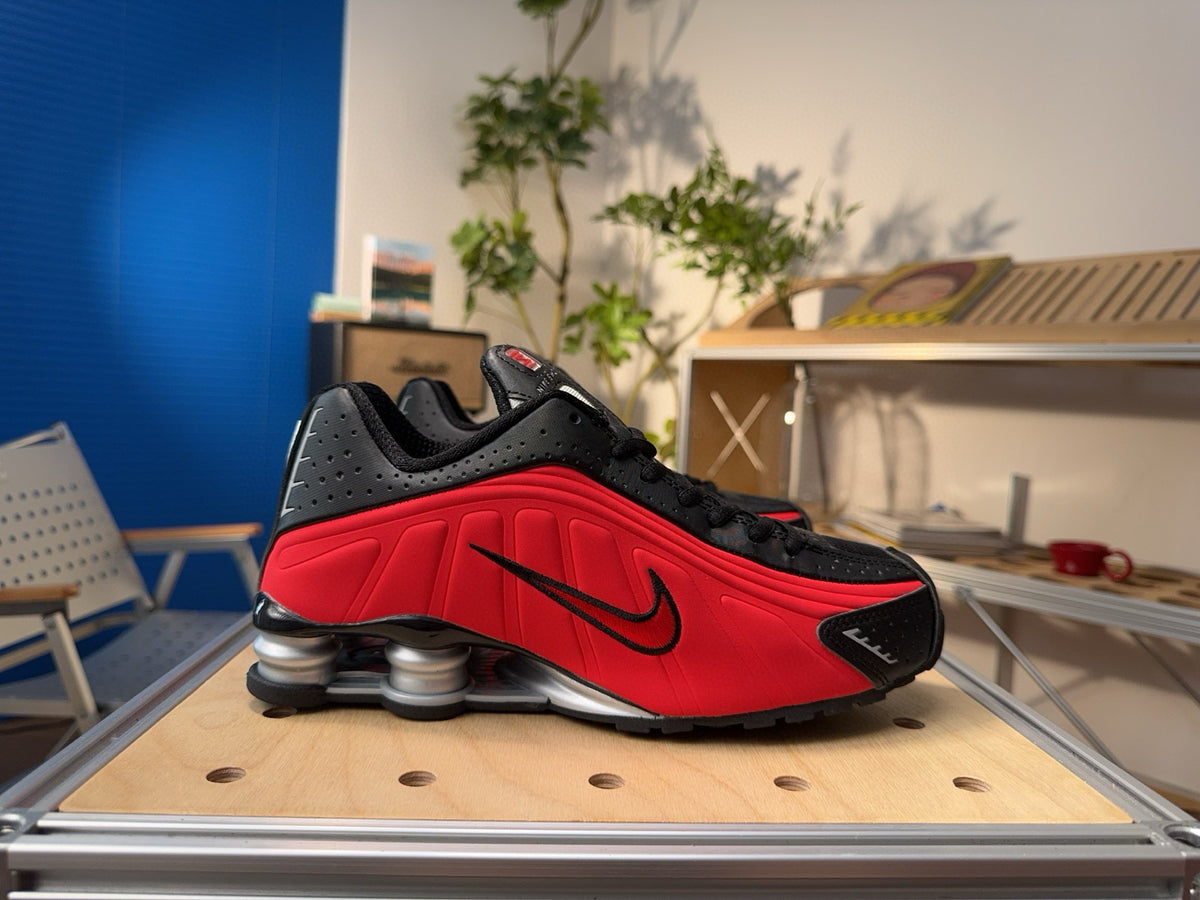 Side view of red and black Nike Shox sneakers with a black Swoosh and heel cushioning columns.