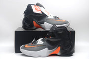 Nike high-top basketball shoes displayed on a black shoebox, showing the side panels, orange accents, and white midsoles.