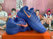 Angled view of two blue Nike sneakers displaying the orange side branding, lace-up construction, and blue outsoles.