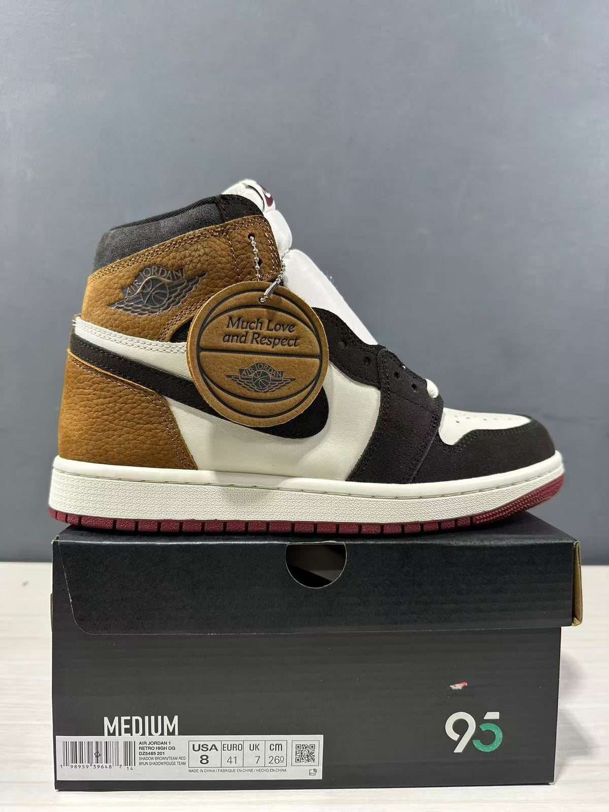 Side view of a Jordan Air Jordan 1 High sneaker in brown, black and white with a burgundy outsole and round hangtag; the box label shows USA 8, EURO 41 and CM 26.