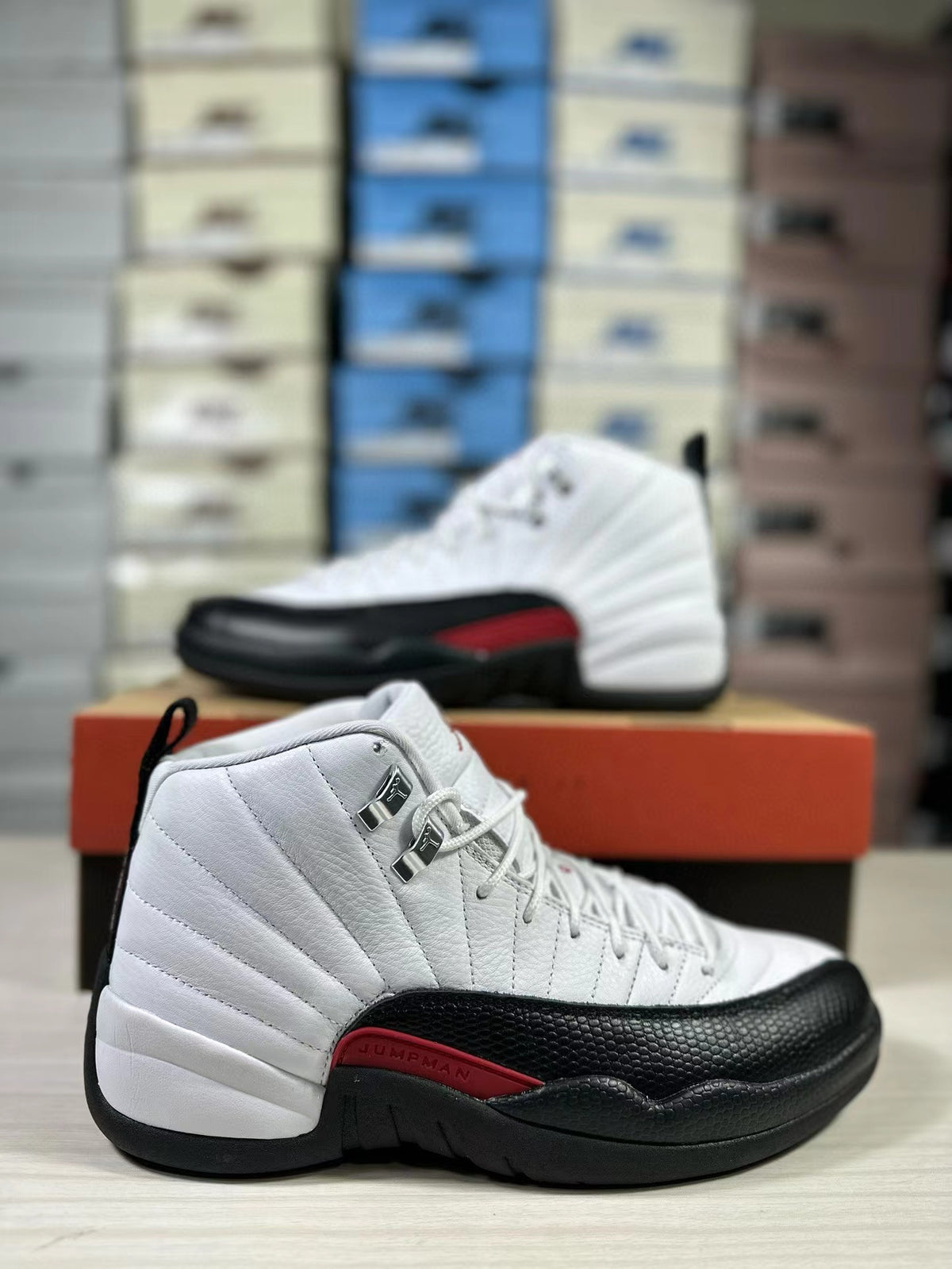 Side view of a white, black and red Jordan Air Jordan 12 high-top sneaker with a matching shoe and stacked boxes in the background.