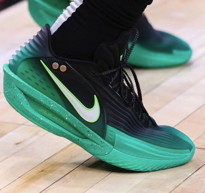 Close-up of a black and green Nike basketball-style sneaker with a white Swoosh, black laces, padded collar, and green speckled sole.