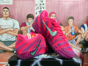 Pair of purple-red Nike basketball sneakers displayed on a black shoebox, showing the patterned upper, green swoosh, and translucent purple outsole.